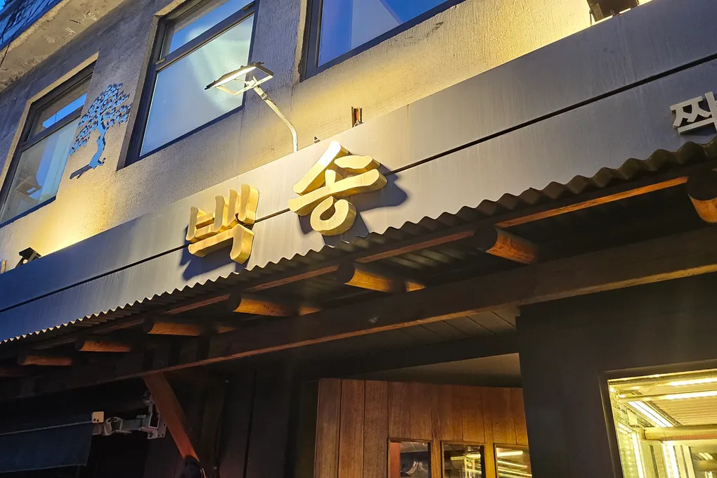 The exterior of Baeksong restaurant in Sindang, Seoul, with a gold sign under a traditional tiled roof at night