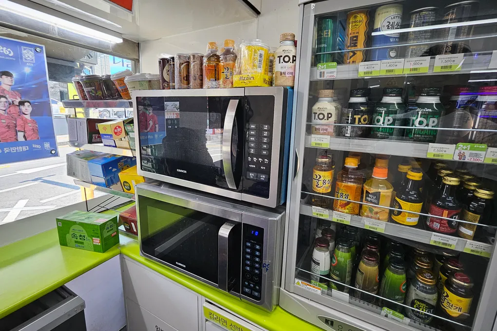 Two microwaves stacked next to a drinks fridge in a Korean convenience store