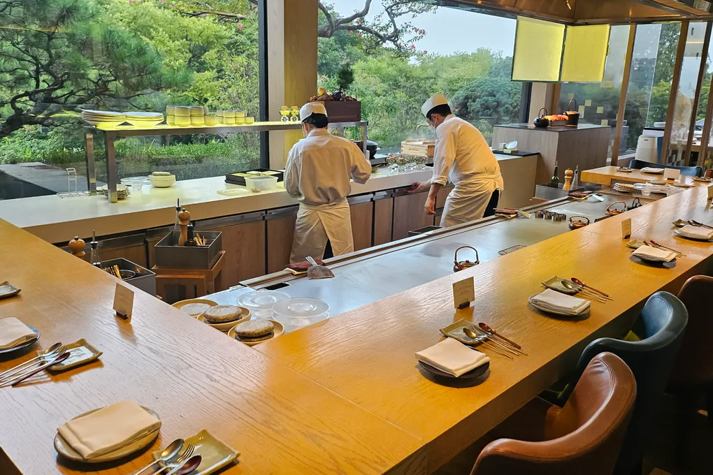 The U-shaped teppanyaki counter at Teppan, Grand Hyatt Seoul, with two chefs in white uniforms working behind the steel griddle and a green garden visible through the windows