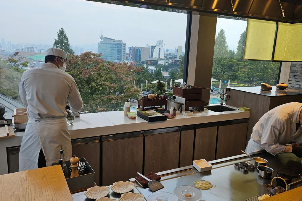 View from the Teppan counter looking out over the Grand Hyatt Seoul outdoor pool, green trees, and the Seoul city skyline, with chefs preparing ingredients in the foreground