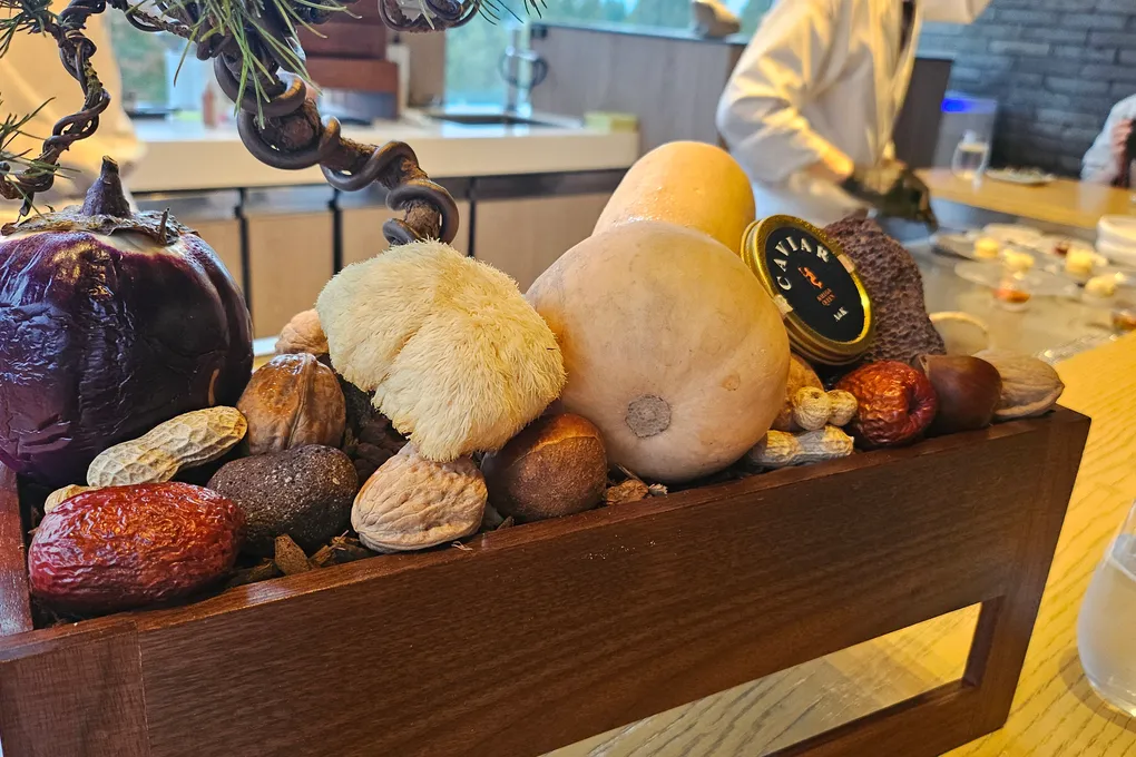 A wooden box on the counter holding autumn produce including a purple eggplant, a white lion's mane mushroom, a squash, chestnuts, walnuts, peanuts and dried dates, with a caviar tin resting on top