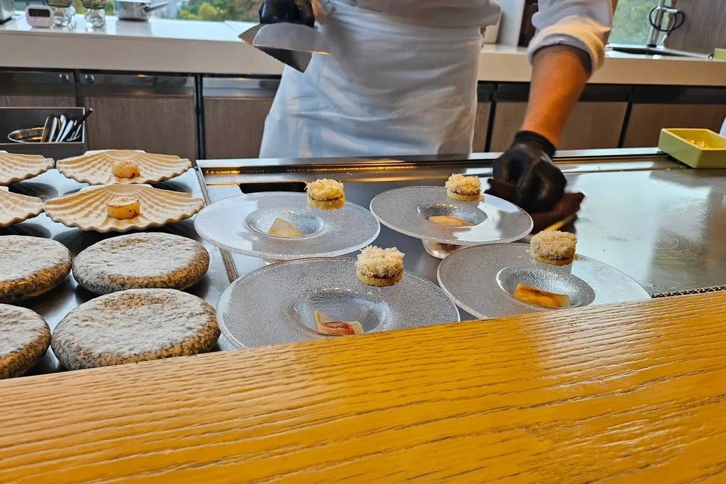 A chef in black gloves plating small bites onto textured white plates and ceramic scallop shells lined up along the edge of the steel griddle