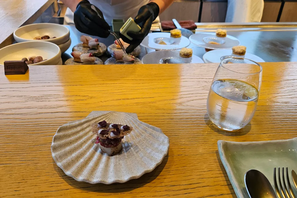 A single small bite topped with thin slices of purple-tinged seafood, served on a cream-colored ceramic scallop shell plate on the wooden counter