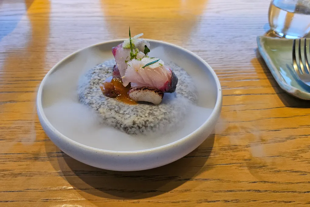 A white ceramic bowl containing raw white fish and mackerel over a speckled grey sesame cream, with a marinated garnish and a small mushroom