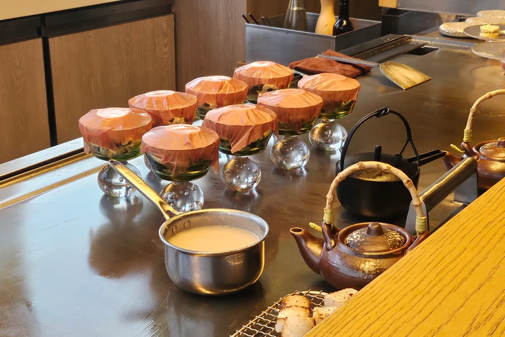 Small glass vessels with tied paper lids arranged on the steel griddle next to copper and cast-iron kettles and a steel pan of pale sauce