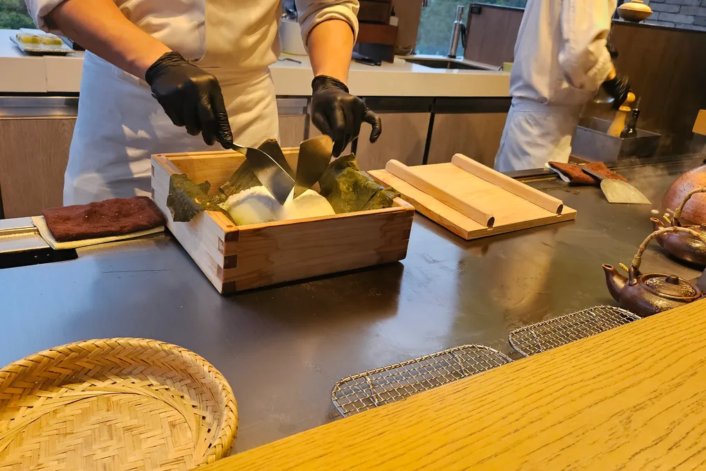 A chef in black gloves opening a wooden box filled with salt and dark green kombu kelp leaves on the teppanyaki griddle, with a slab of beef resting to the left