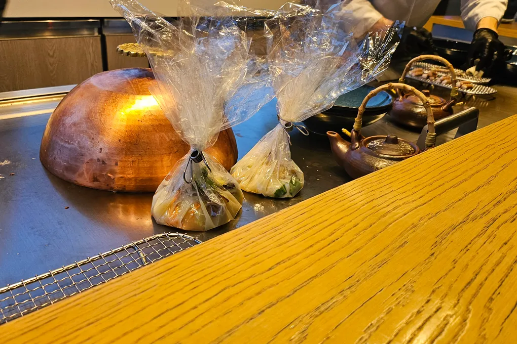 Two cellophane-wrapped parcels tied with clips steaming on the griddle beside a large copper dome cover