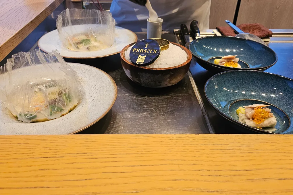 A tin of Perseus caviar set in a bowl of crushed ice on the griddle, next to two deep blue bowls holding white fish topped with caviar and roe, with cellophane-wrapped parcels on plates alongside
