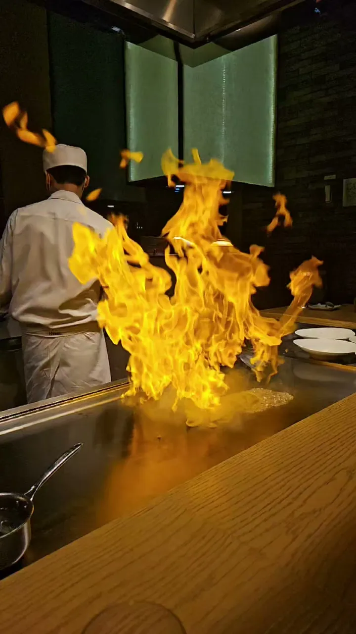 A tall column of orange flame rising from the teppanyaki griddle at Teppan, Grand Hyatt Seoul, with a chef in white standing behind it in the darkened restaurant