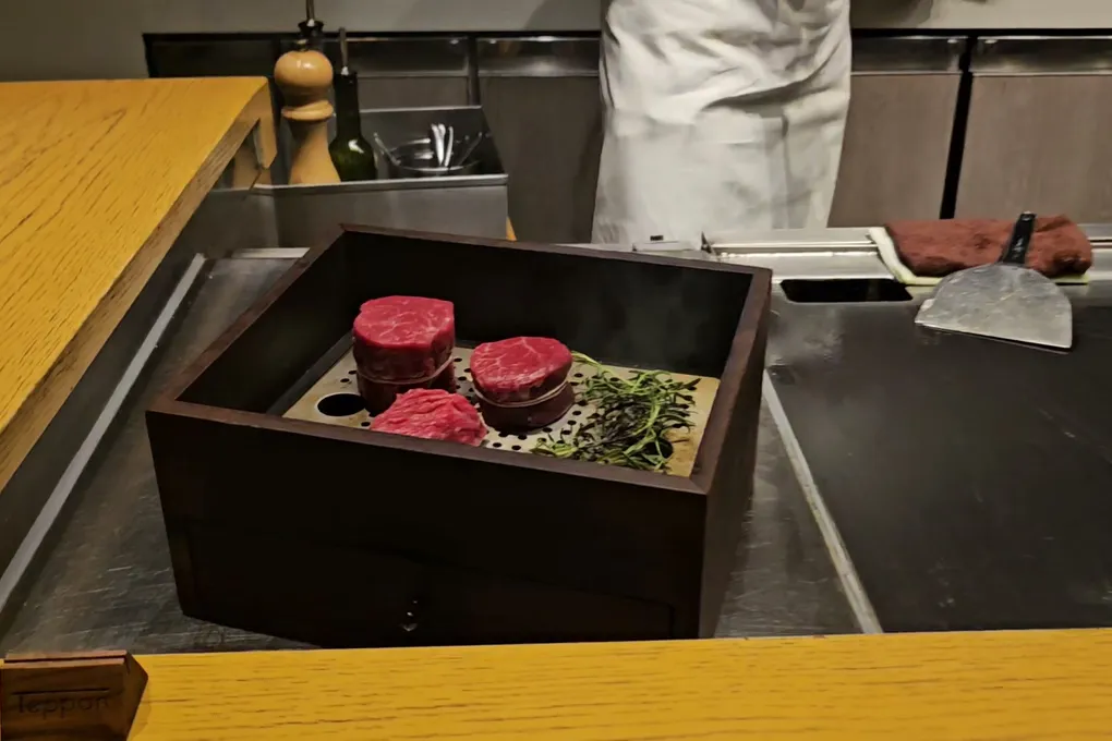 Three thick raw beef medallions displayed in a dark wooden box on a bed of green herbs with smoke rising, sitting on the griddle