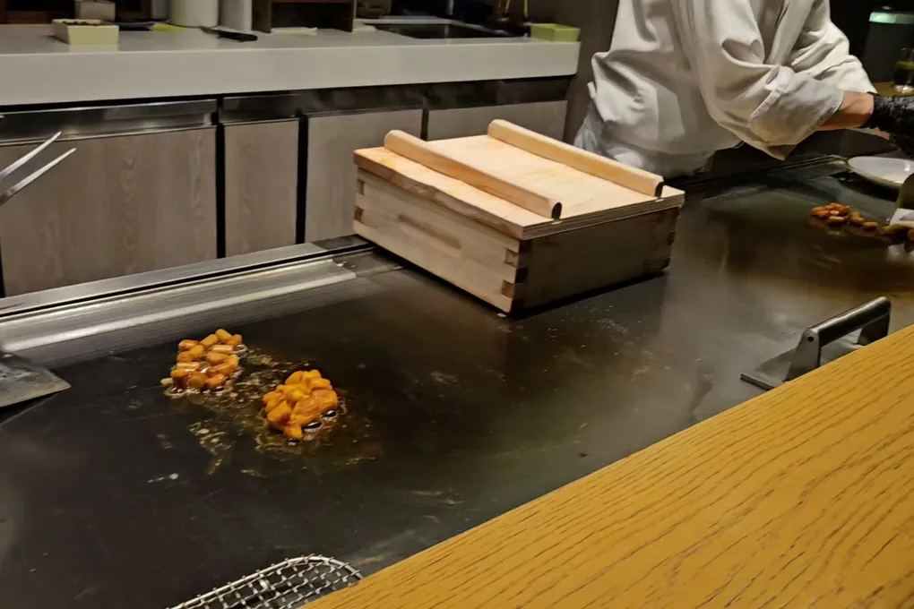 Lobes of orange sea urchin searing directly on the steel teppanyaki griddle at night, with a wooden box and skewers nearby